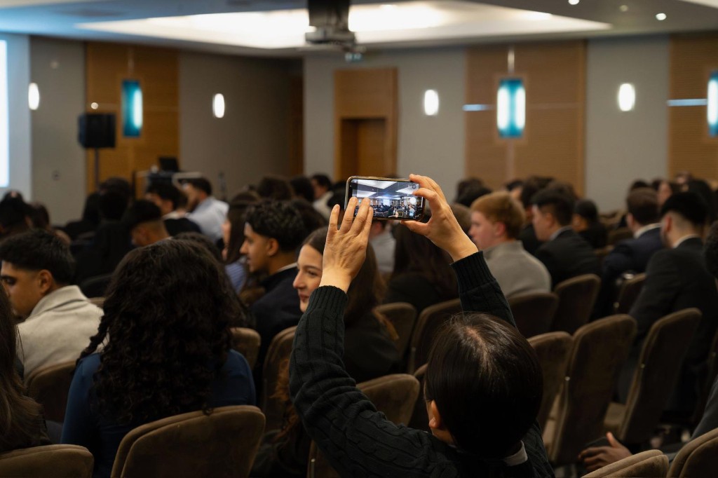 Conference hall: attendee photographing the session on a phone, rows of professionals seated in a modern venue